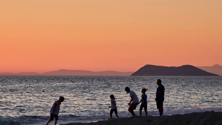 A happy family with young children walking hand-in-hand on a sunny beach, enjoying their family trip to Greece.