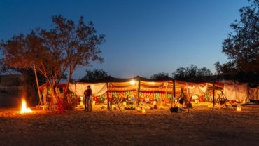 A traditional Berber desert camp for camping in the Sahara, with tents lit up under a starry night sky and a warm campfire in the foreground.