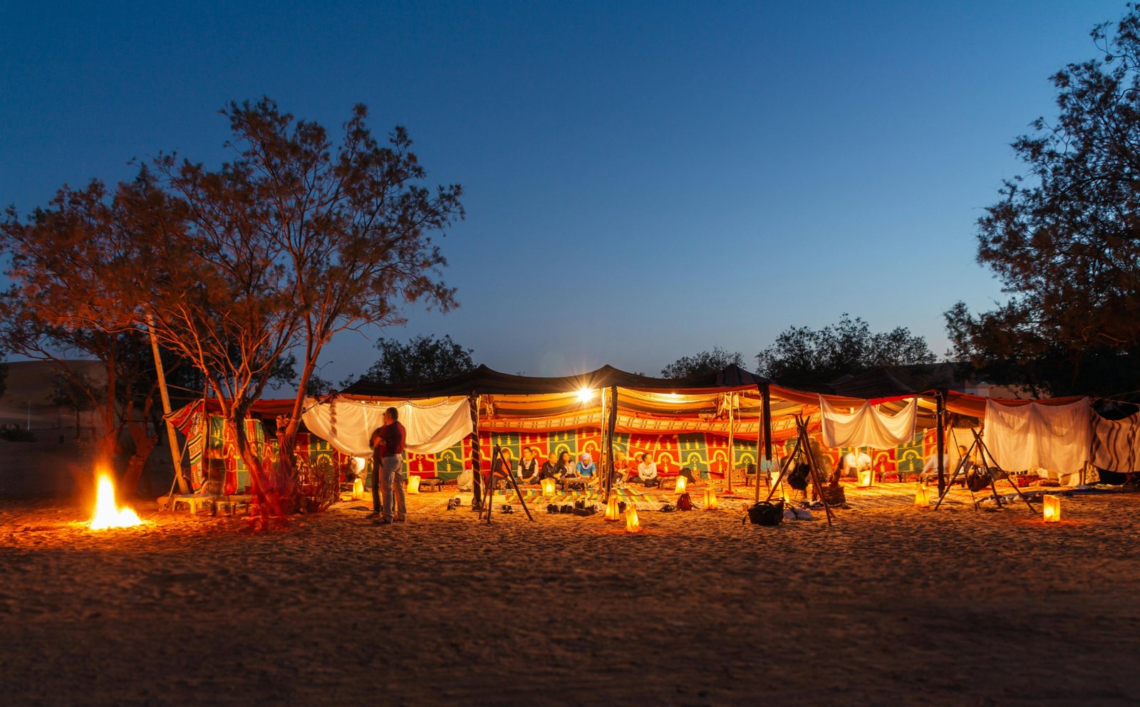 A traditional Berber desert camp for camping in the Sahara, with tents lit up under a starry night sky and a warm campfire in the foreground.