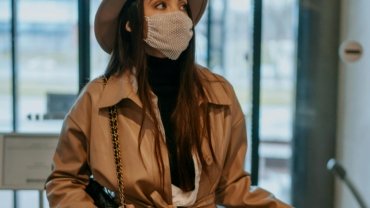 A stylish traveler, possibly a woman, effortlessly carrying a 21 cm compact travel hat box with her luggage at an airport, symbolizing protection for hats like fascinators or fedoras and adding a touch of vintage elegance to her journey.