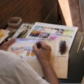 A close-up of an artist's hands using a portable travel watercolor set to paint a beautiful landscape outdoors, emphasizing the convenience and inspiration for creative adventurers.
