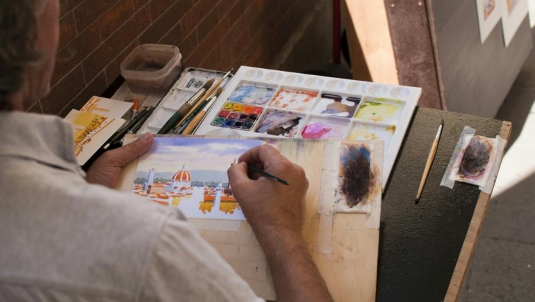 A close-up of an artist's hands using a portable travel watercolor set to paint a beautiful landscape outdoors, emphasizing the convenience and inspiration for creative adventurers.