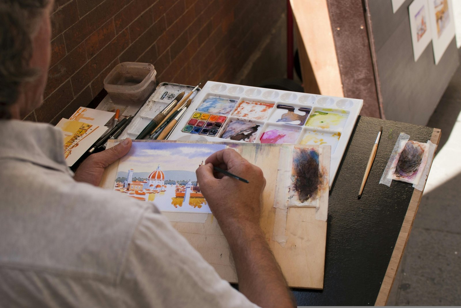 A close-up of an artist's hands using a portable travel watercolor set to paint a beautiful landscape outdoors, emphasizing the convenience and inspiration for creative adventurers.