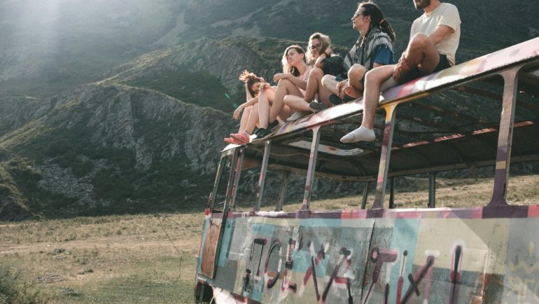 Diverse young backpackers with backpacks smiling on a hop-on hop-off 'traveller bus' against a beautiful natural backdrop, symbolizing budget-friendly, social, and adventurous travel exploration.
