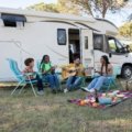 A modern travel trailer with a gray slide-out awning (slide topper) extended, providing essential protection from sun, rain, and debris for the RV's slide-out in a scenic outdoor camping environment.