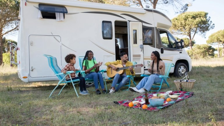 A modern travel trailer with a gray slide-out awning (slide topper) extended, providing essential protection from sun, rain, and debris for the RV's slide-out in a scenic outdoor camping environment.
