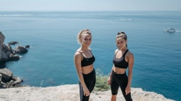 Two young women, resembling travel fitness influencers and sisters, joyfully exploring a vibrant travel destination, carrying reusable water bottles and embodying a healthy, active wellness lifestyle on the road. This image illustrates tips for maintaining health and fitness while traveling.