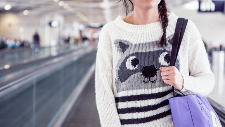 Stylish woman effortlessly walking through a modern airport terminal with a sleek travel tote bag secured to her rolling luggage handle via a trolley sleeve, illustrating hands-free and organized travel convenience.