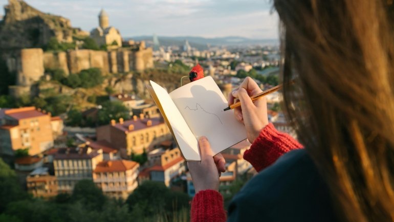 Close-up of hands writing in a durable travel notebook with a pen, documenting travel memories and experiences against a blurred scenic landscape, promoting mindful journaling on adventure.