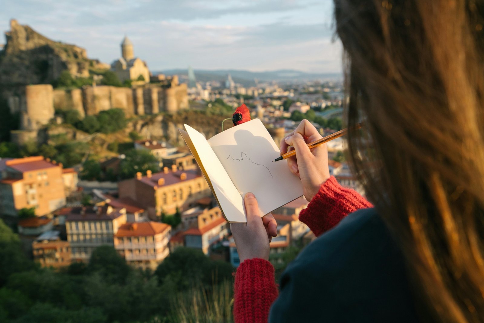 Close-up of hands writing in a durable travel notebook with a pen, documenting travel memories and experiences against a blurred scenic landscape, promoting mindful journaling on adventure.