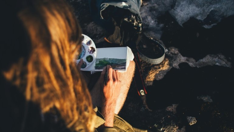 Artist using a portable watercolor travel set and sketchbook to paint a vibrant outdoor landscape, perfect for capturing travel memories on the go.