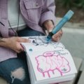 Close-up of hands sketching in a travel journal or sketchbook with a fineliner pen, overlooking a scenic European city street or a cozy cafe, illustrating the mindful art of travel drawing and creating personal souvenirs.