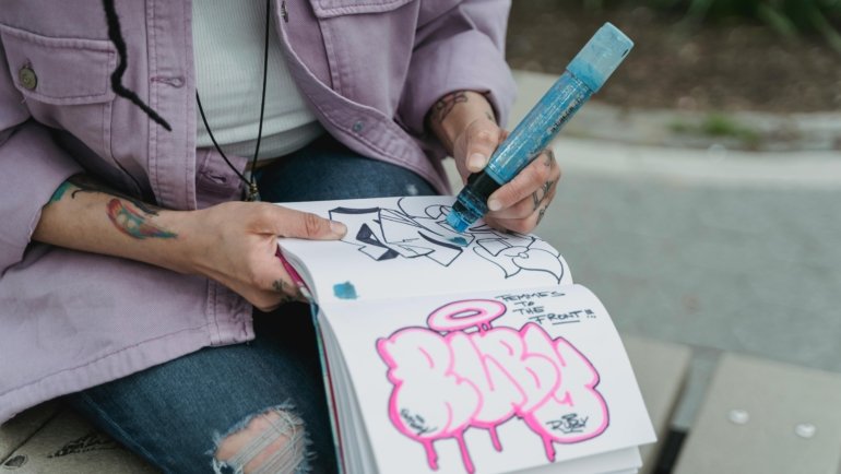 Close-up of hands sketching in a travel journal or sketchbook with a fineliner pen, overlooking a scenic European city street or a cozy cafe, illustrating the mindful art of travel drawing and creating personal souvenirs.