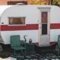A modern destination travel trailer featuring a spacious, residential-style interior with high ceilings and a sliding glass door opening to a comfortable patio, suggesting long-term, home-like living in a serene campsite.