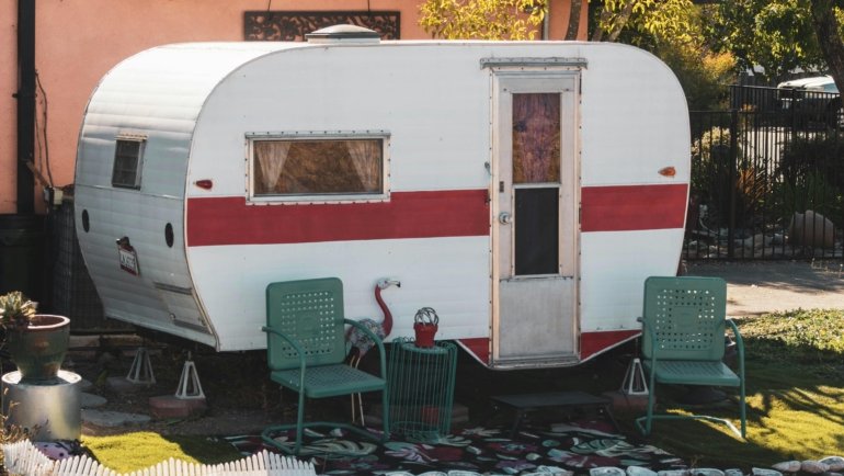 A modern destination travel trailer featuring a spacious, residential-style interior with high ceilings and a sliding glass door opening to a comfortable patio, suggesting long-term, home-like living in a serene campsite.
