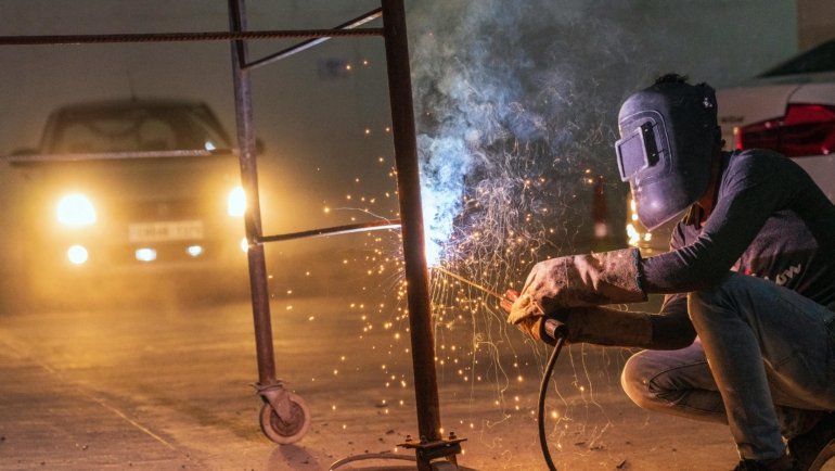 Professional traveling welder in full gear creating bright sparks while working on a large industrial pipeline or construction site, representing skilled journeyman welding.