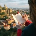 Traveler's hands sketching a scene in a personal travel journal with a pen, illustrating the mindful practice of traveling drawing and creating unique souvenirs.