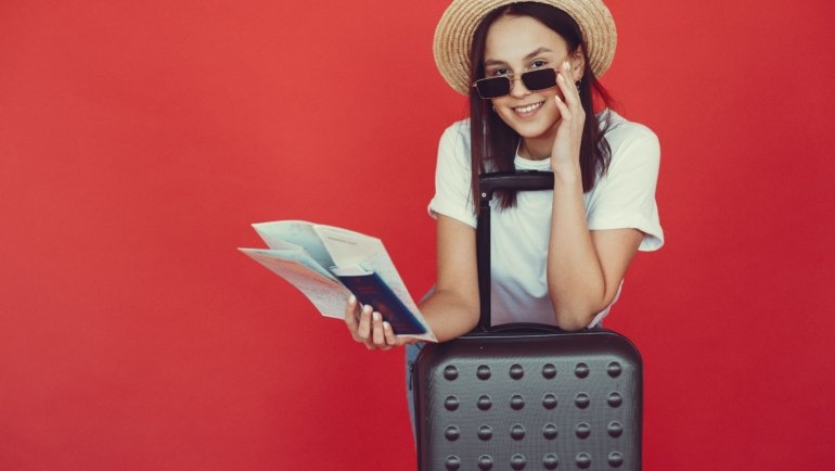Smiling traveler holding round-trip plane ticket with luggage at airport, representing convenient and affordable travel planning